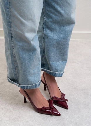 Bordo LUCINDA Slingback-Pumps aus glänzendem Lackleder mit spitzer Vorderkappe, mittelhohem schmalen Absatz und verstellbarem Slingback-Riemen. Dekorative Schleife auf der Vorderseite.